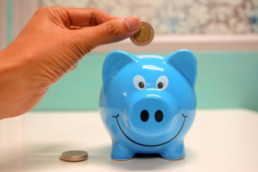 This picture shows a blue piggy bank, the piggy bank has a big smile and a hand is placing money into the slot