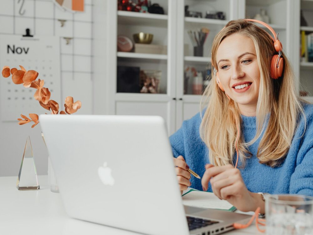 This picture shows a young woman with headphones on watching a laptop screen whilst sat at their desk