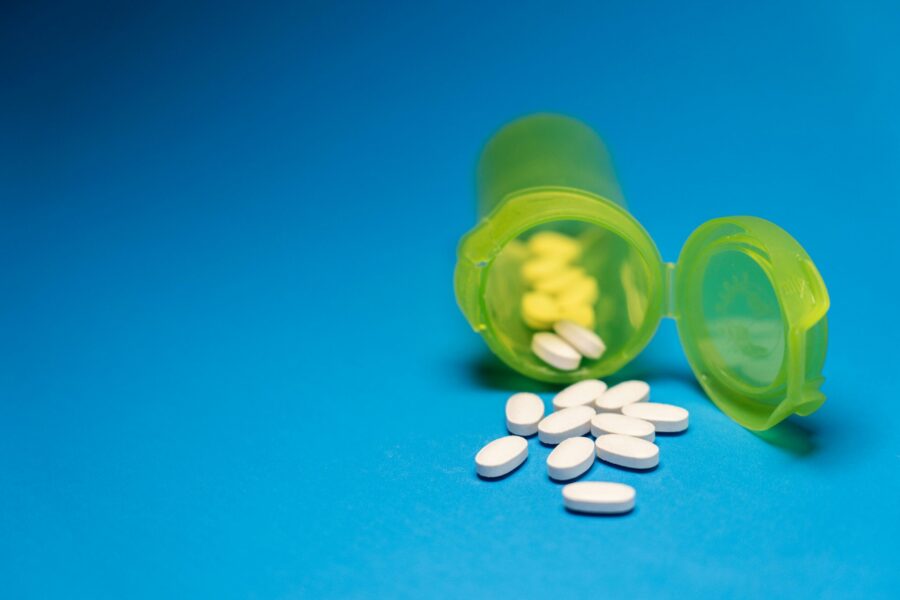 This shows a blue background and a green pill container, the container is on its side with the top one and little white pills have spilled out.